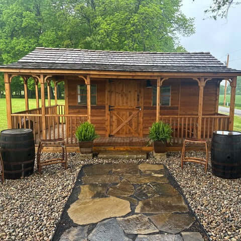 Wooden shed with a covered porch, double doors, and windows, set in a grassy outdoor area with trees in the background.