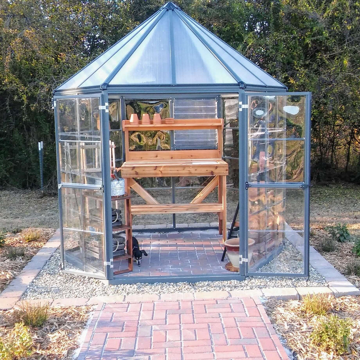Wooden potting bench with upper and lower shelves and sink, shown inside a glass greenhouse with open doors.